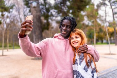 Canlı bir parkta neşeli bir selfie çekerken, gülümseyen ve sarılan genç çift, dışarıda aşklarını ve mutluluklarını yakalıyorlar.