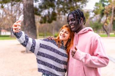 Parkta güneşli bir günün tadını çıkaran mutlu çoklu etnik çift coşkulu bir şekilde selfie çekiyorlar.