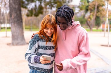 Parkta neşeli bir anı paylaşan iki yetişkin, akıllı telefona gülümserken arkadaşlığın ve doğanın tadını çıkarıyor.