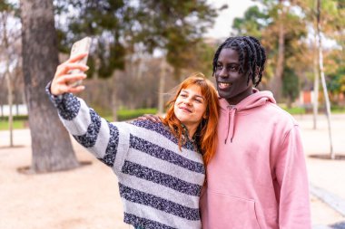 Canlı bir parkta gülümseyen ve selfie çeken ırklar arası bir çift, yeşillik ve doğa ile çevriliyken birbirlerinin arkadaşlığından keyif alıyorlar.