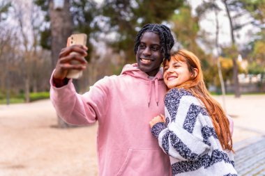 Canlı bir parkta akıllı bir telefonla selfie çekerken gülümseyen çok ırklı mutlu bir çift açık havada eğleniyor.