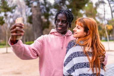 Genç yetişkinler bir park ortamında selfie çekerek neşeli bir anı yakalıyorlar. Gülümseyerek ve dışarıda eğlenerek, teknolojiyi ve arkadaşlığı kucaklarlar.