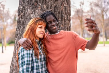 Dışarıda selfie çekerken birlikte gülümseyen iki genç yetişkin park ortamında dostluk ve neşe yakalıyorlar.