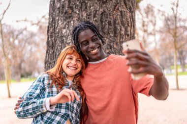 Mutlu çoklu etnik çiftler gülümsüyor ve canlı bir parkta akıllı telefonlarıyla selfie çekiyorlar. Açık havada eğleniyorlar.
