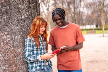 İki genç yetişkin parkta akıllı telefonlarına bakarken bir anı paylaşıyorlar. Gülümsüyorlar ve cihazlarındaki içerikle meşgul görünüyorlar.