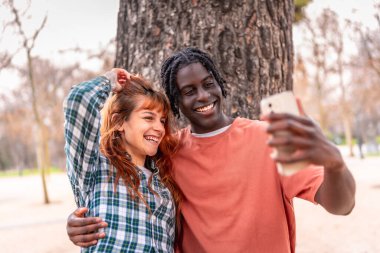 Mutlu çoklu etnik çift gülüyor ve canlı bir parkta selfie çekiyor, ağaçların altında eğleniyorlar.