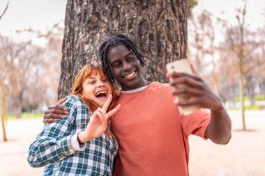 Canlı bir parkta gülümseyen ve poz veren iki genç arkadaş, neşeyle barış işaretleri yapıyor ve birlikte mutlu anları yakalıyorlar.