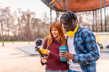 Parkta video çekerken gülümseyen ve telefonlarına bakan iki genç içerik yaratıcısı