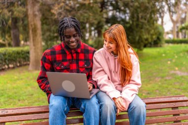 İki genç öğrenci parkta bir bankta oturuyor, dizüstü bilgisayara bakıyor ve bir projede işbirliği yapıyor, temiz havanın ve doğal çevrenin tadını çıkarıyorlar.