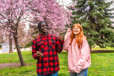 Irklar arası genç çift bahar mevsiminde çiçek açan kiraz ağaçlarıyla dolu parkta el ele tutuşup gülüyorlar.