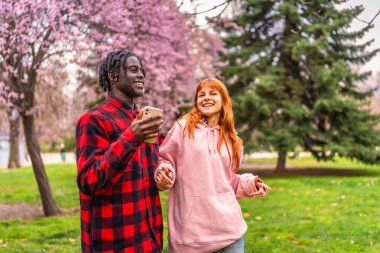 Genç, çok ırklı bir çift, baharda parkta birlikte yürüyor, çiçek açan ağaçların tadını çıkarıyor ve akıllı telefon kullanıyor.