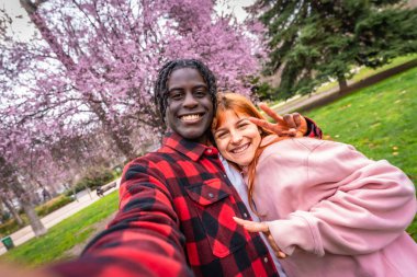 Mutlu çoklu etnik çift pembe çiçeklerle dolu bir parkta selfie çekiyor barış işaretleri yapıyor ve kameraya gülümsüyor.