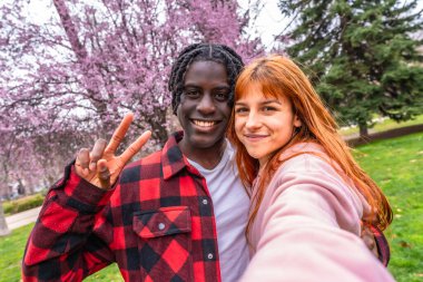 Mutlu çoklu etnik çift gülümseyip ağaçların altında selfie çekiyor, güzel bir bahar gününün tadını çıkarırken neşe içinde barış işaretleri yapıyor.