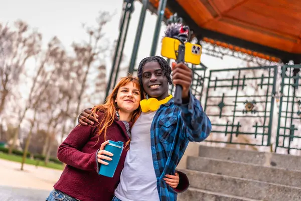 İki genç içerik yaratıcısı parkta video çekiyor, akıllı bir telefon ve telle çekim yapıyor, gülümsüyor ve kameraya bakıyor.