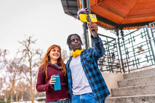 İki genç içerik yaratıcısı bir şehir parkında mikrofonlu bir akıllı telefon kullanarak video kaydediyor.