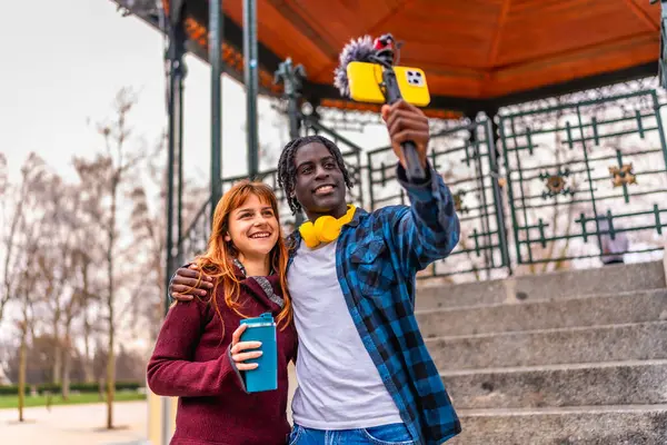 İki genç içerik yaratıcısı bir şehir parkında bir video blogu kaydediyor, mikrofonlu bir akıllı telefon kullanıyorlar.