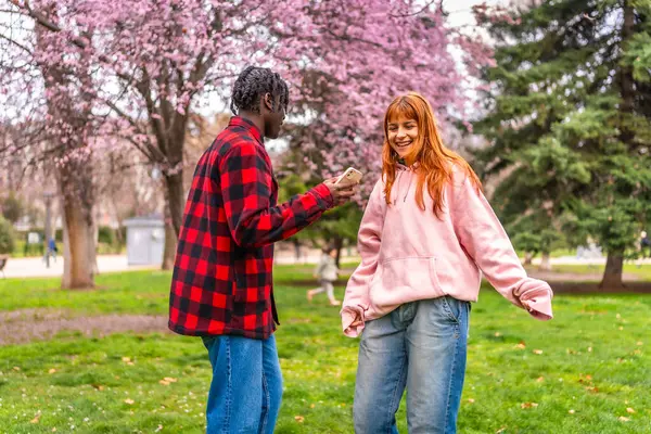 Genç adam, kız arkadaşının neşeli anlarını canlı bir parkta canlı bir telefonla yakalıyor. Baharda çiçek açan çiçeklerle çevrili.