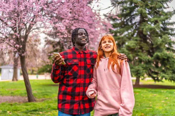 Çiçek açan ağaçlarla parkta yürüyen ırklar arası genç bir çift. Adamın kolu kadının omuzlarında ve elinde akıllı bir telefon var.