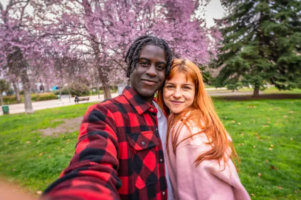 Parkta çiçek açan kiraz ağaçlarının altında selfie çeken, bahar mevsiminin tadını çıkaran çok ırklı mutlu bir çift.