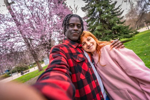 Parktaki kiraz ağaçlarının altında gülümseyen ve selfie çeken mutlu çoklu etnik çift, güzel bir bahar gününün tadını çıkarıyor.