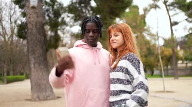 Çoklu etnik çeşitlilikte genç bir çift selfie anları yakalıyor, gülüyor ve kaygısız bağlantıların keyfini çıkarırken manzaralı şehir parkı ortamında samimi duyguları paylaşıyorlar.