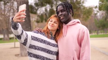 Genç, çok ırklı bir çift, güneşli şehir parkında birlikte vakit geçirirken akıllı telefon özçekimleri yapıyor, neşe ve bağlarını ifade ediyorlar.
