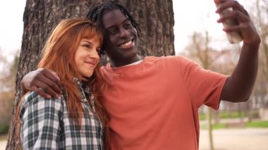 Çok ırklı genç bir çift, neşeli selfie çekiyorlar. Neşeli anları ve şefkati paylaşıyorlar. Açık hava parkının doğal güneş ışığıyla tadını çıkarıyorlar.