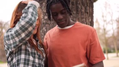 Genç çift, yeşil ağaçlarla çevrili güneşli şehir parkında akıllı telefonlara bakarken neşeli anları paylaşıyor.