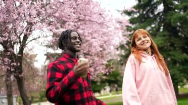 Çok ırklı genç çift, canlı bahar mevsiminde güneşli şehir parkında çiçek açan kiraz çiçeklerinin arasında birlikte yürüyor ve samimi sohbetler ediyorlar.