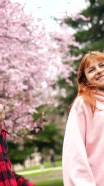 Genç, çok ırklı bir çift birlikte yürüyor, canlı bir bahar öğleden sonrası boyunca manzaralı parkta pembe kiraz çiçekleri altında samimi sohbetler ediyorlar.