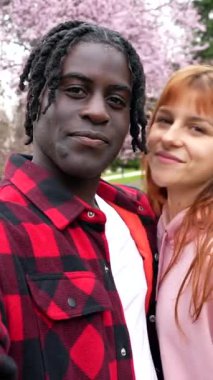Genç, çok ırklı bir çift romantik bir anı yakalıyor. Çiçek açan kiraz ağaçlarının altında selfie çekiyorlar. Etrafı manzaralı parkta pembe yapraklarla çevrili.