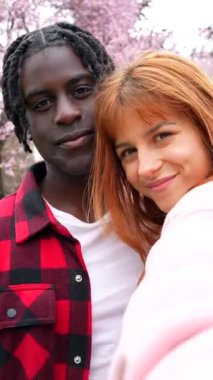 Genç çok ırklı çift, çiçek açan kiraz çiçeklerinin altında romantik bir anın tadını çıkarır. Aşklarını neşe dolu selfielerle yakalarlar.