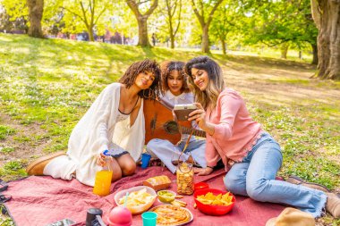 Üç kadın parkta neşeli bir piknik sırasında klasik bir kamerayla selfie çekiyor. Atıştırmalık ve içeceklerin tadını çıkarıyorlar.
