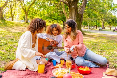 Üç farklı kadın battaniyede dinleniyor, abur cuburlardan hoşlanıyor, müzik çalıyor ve parkta güneşli bir piknik sırasında fotoğraf çekiyor.
