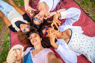 Parkta bir battaniyeye uzanmış dört neşeli genç kadın el hareketi yapıyor ve selfie çekiyor.