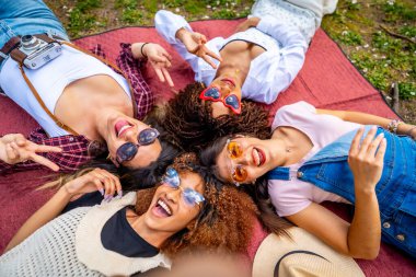 Dört çok ırklı bayan arkadaş parkta bir battaniyenin üzerine uzanmış selfie çekiyor ve barış işaretleri gösteriyorlar.