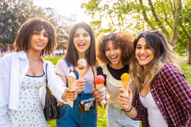 Dört gülümseyen, etnik çeşitlilikte kadın güneşli bir yaz gününde parkta dinlenirken dondurma yiyip arkadaşlığı ve eğlenceyi kutluyor.
