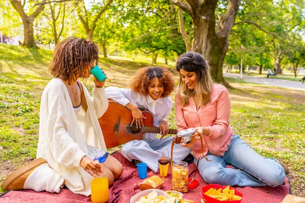 Üç bayan arkadaş parkta piknik yapıyor, gitar çalıyor, fotoğraf çekiyor ve güneşli bir günde atıştırmalık ve içeceklerden keyif alıyorlar.