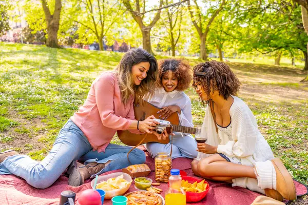 Üç arkadaş parkta pikniğin tadını çıkarıyor, eski bir kamerayla fotoğraf çekiyor, gitar çalıyor ve güneşli bir günde yemek ve içecekleri paylaşıyor.