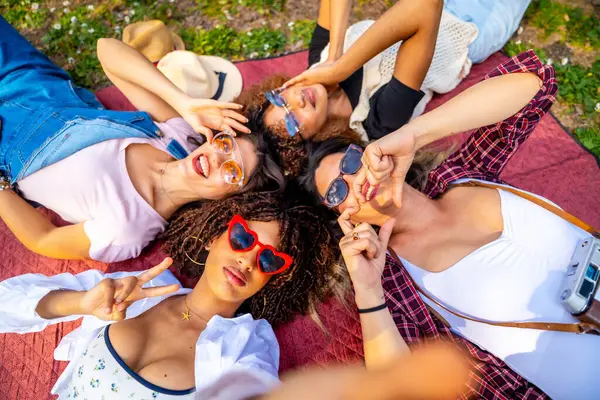 Güneş gözlüğü takan dört neşeli genç kadın parkta bir battaniyeye uzanmış selfie çekiyor ve komik suratlar yapıyor.