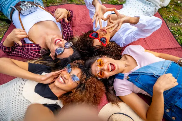 Dört genç kadın battaniyeye uzanıp elleriyle kalp şekli oluşturuyorlar. Selfie çekiyorlar ve gülümsüyorlar.