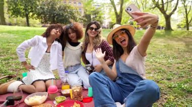 Genç bayan arkadaşlar güneşli parkta piknik yaparken birbirlerine bağlanıyorlar, selfie çekiyorlar, yeşil çimenlerde dinlenirken neşe ve dostluk anlarını paylaşıyorlar.