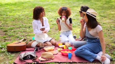 Parkta neşeli bir pikniği paylaşan arkadaşlar, güneşli bir günde yemeğin, içkinin ve sohbetin tadını çıkarıyorlar.