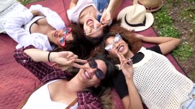 Piknik battaniyesinde uzanan, güneş gözlüğü takan ve güneşli parkın keyfini çıkarırken komik ifadeler kullanan genç kadınlar.