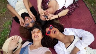 Parkta battaniyeye uzanmış, arkadaşlıklarının tadını çıkarırken elleriyle kalp yapan üç genç kadın.