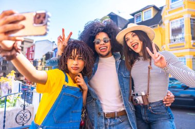 Üç mutlu genç turist akıllı telefonlarıyla selfie çekiyorlar ve renkli bir şehri ziyaret ederken barış işareti yapıyorlar.