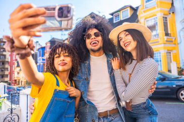 Arka planda renkli binalarla selfie çeken üç mutlu turist, tatillerinin tadını çıkarıyor ve hareketli bir şehri keşfediyorlar.