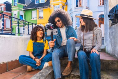 Gülümseyen arkadaşlar renkli bir mahallede mikrofonlu akıllı telefon kullanarak video bloğu oluşturuyorlar.