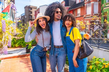 Üç genç turist ellerinde kamerayla sarılıp gülümsüyor, renkli bir şehir ortamında tatilin tadını çıkarıyorlar.