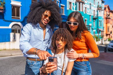 Üç genç turist renkli bir sokakta akıllı telefon kullanıyor, sosyal medya içeriğini paylaşıyor ve tatillerinin tadını çıkarıyorlar.